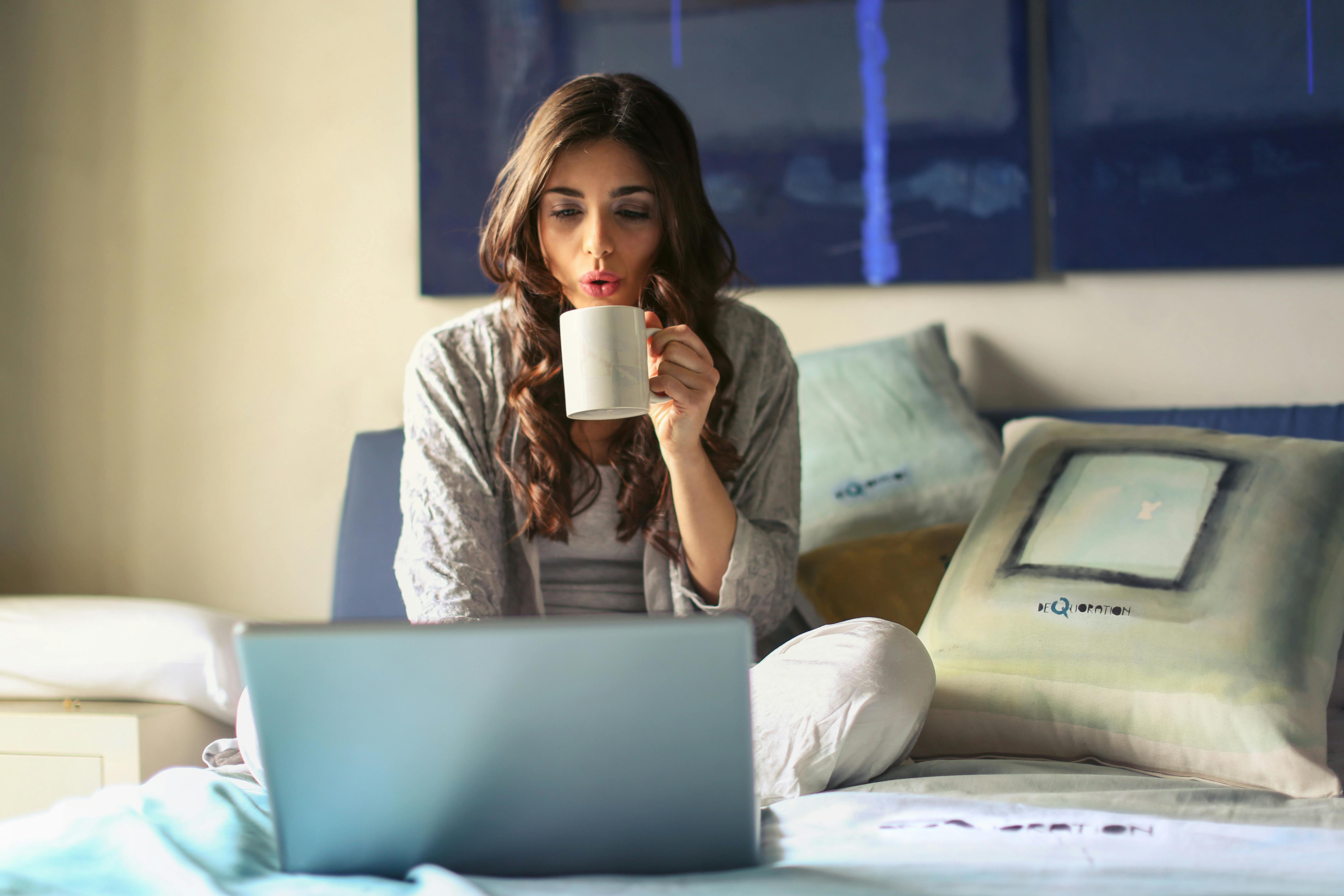 Woman on her laptop in bed while drinking coffee in a mug