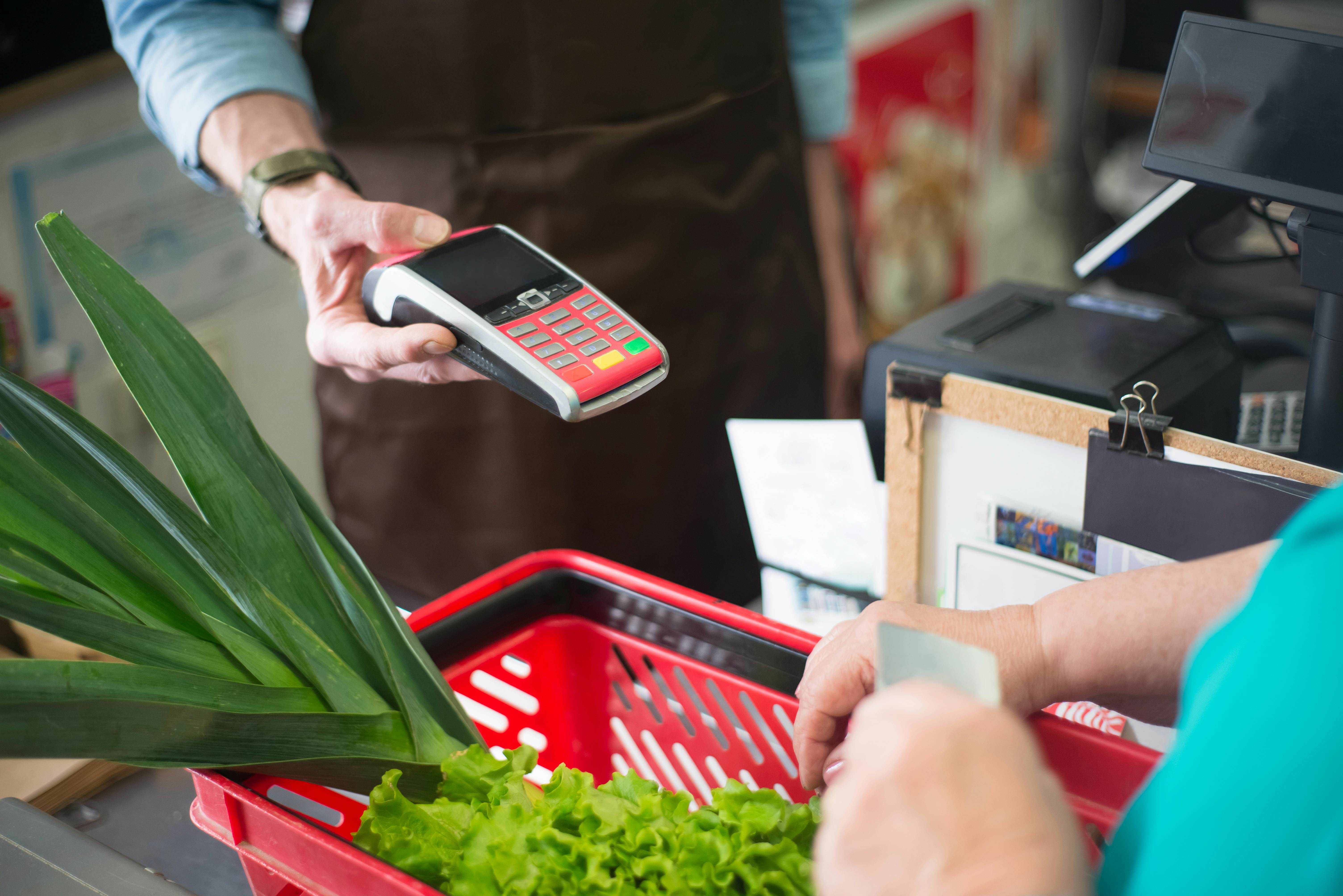 A man holds out a credit card reader to a woman who holds credit cards above a basket of vegetables.