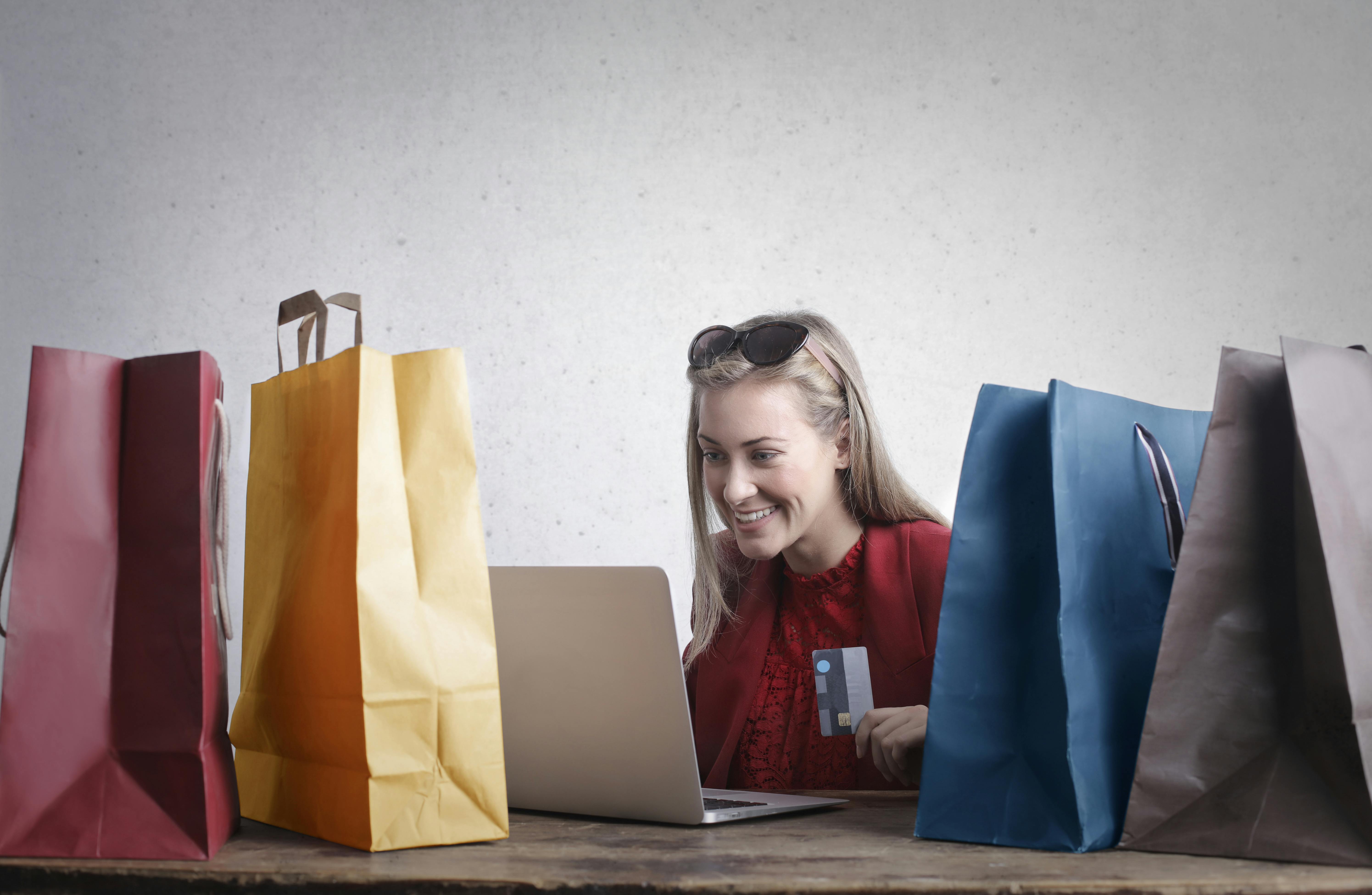 A woman holds up a credit card as she looks at her laptop screen. There are shopping bags on either side of her.
