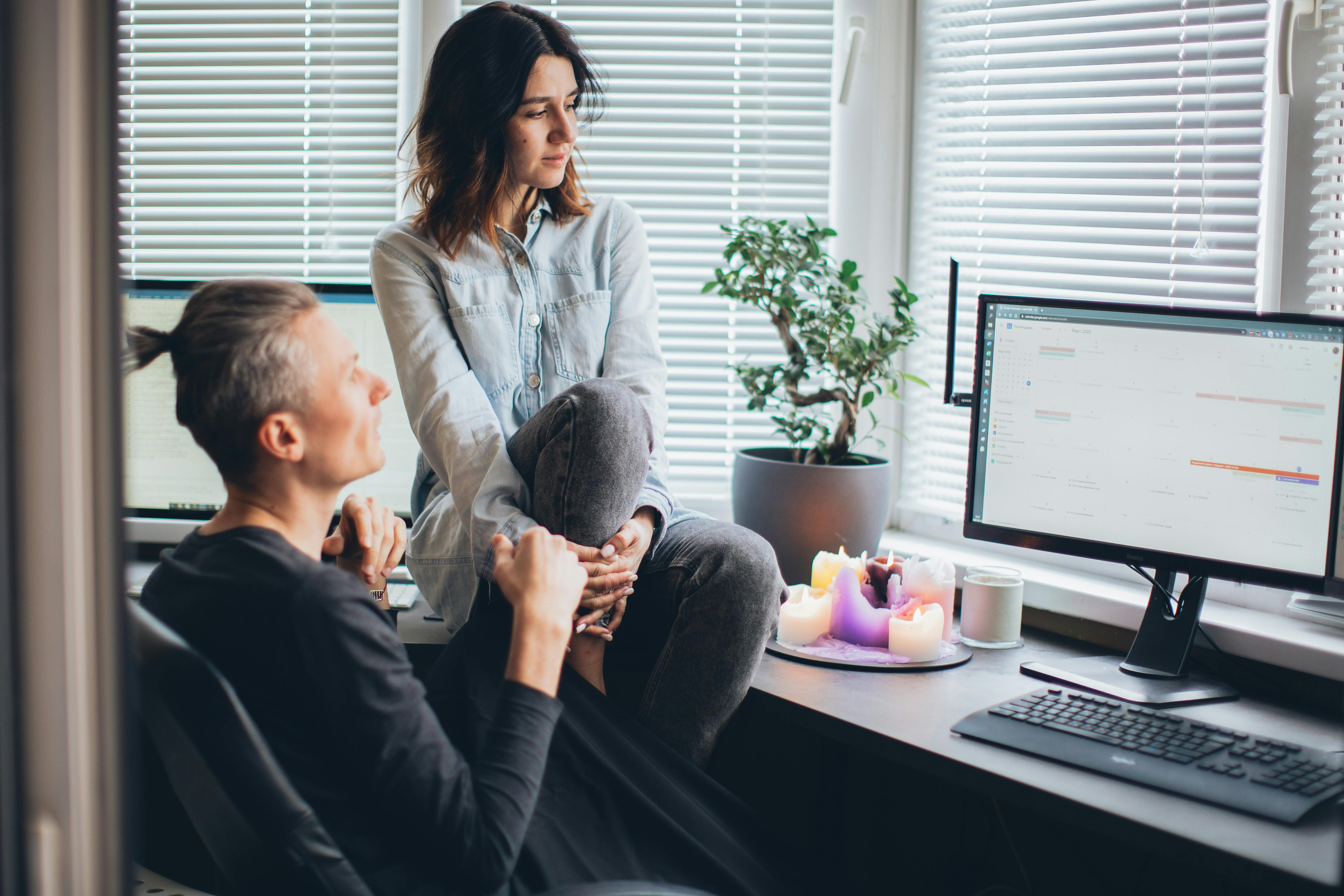 A woman sits on a desk, looking at a computer monitor while a man sits in the desk chair.