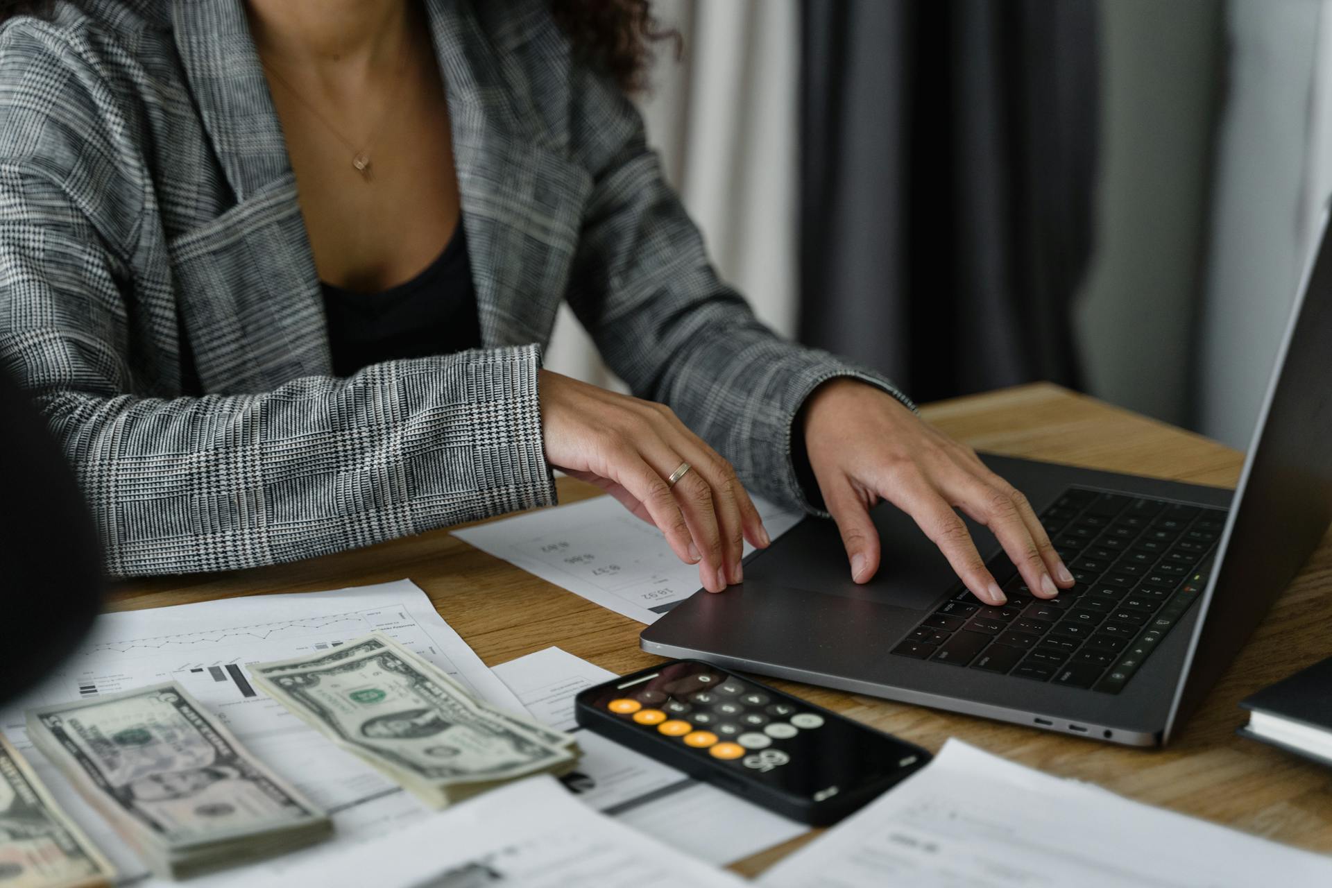 A woman types on a laptop with a calculator and small stacks of cash next to her.